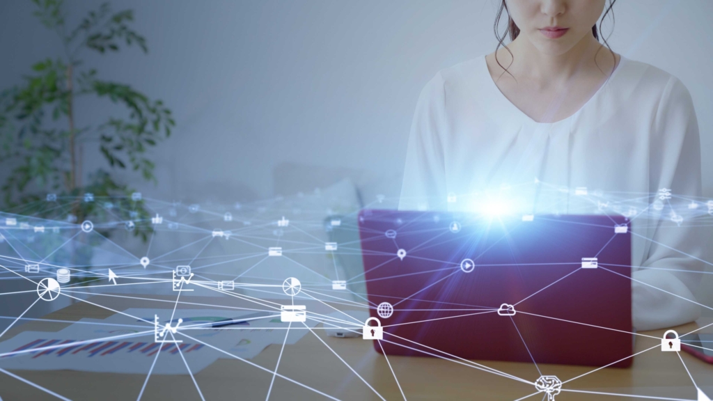 Patch Deployment Automation. A woman sits at a desk using a tablet, partially obscured by a bright digital overlay of interconnected icons such as locks, clouds, and gears. The imagery represents digital security and network communication.